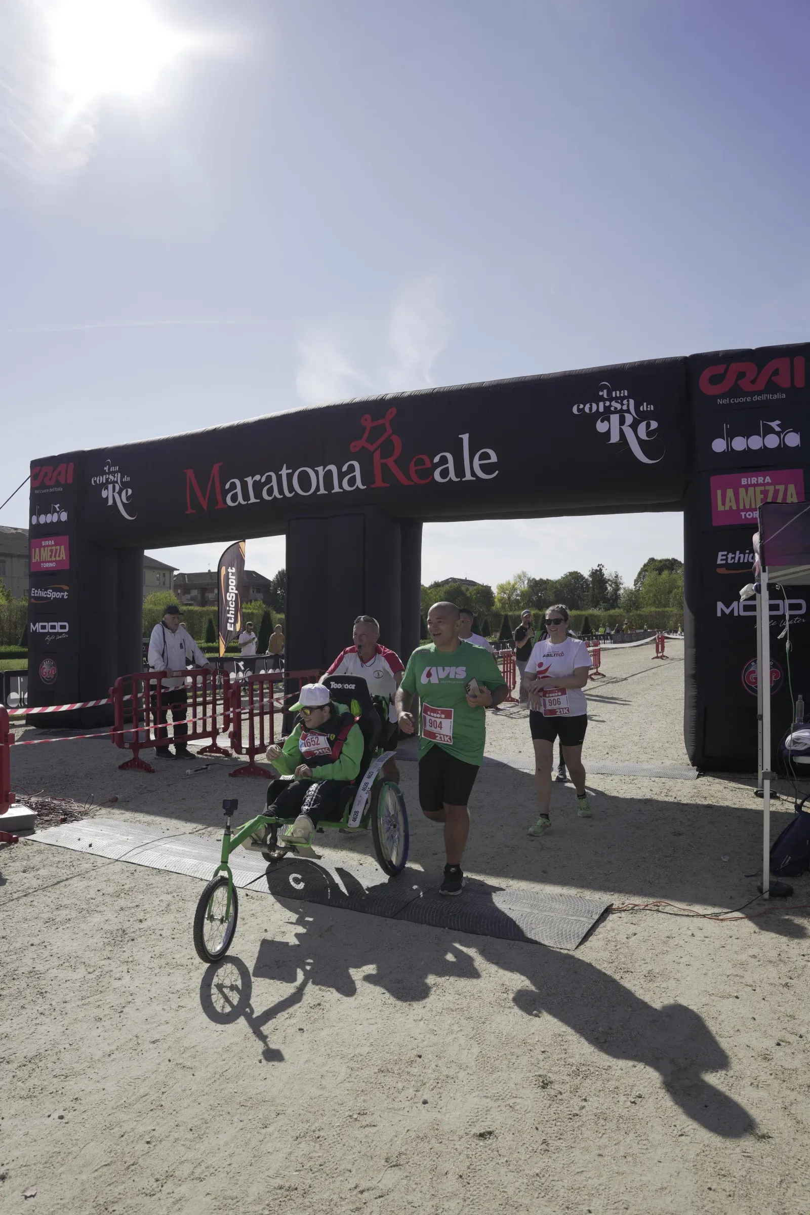 Un ragazzo sulla joelette taglia il traguardo della mezza maratona spinto dai volontari, sotto l'arco della Maratona Reale
