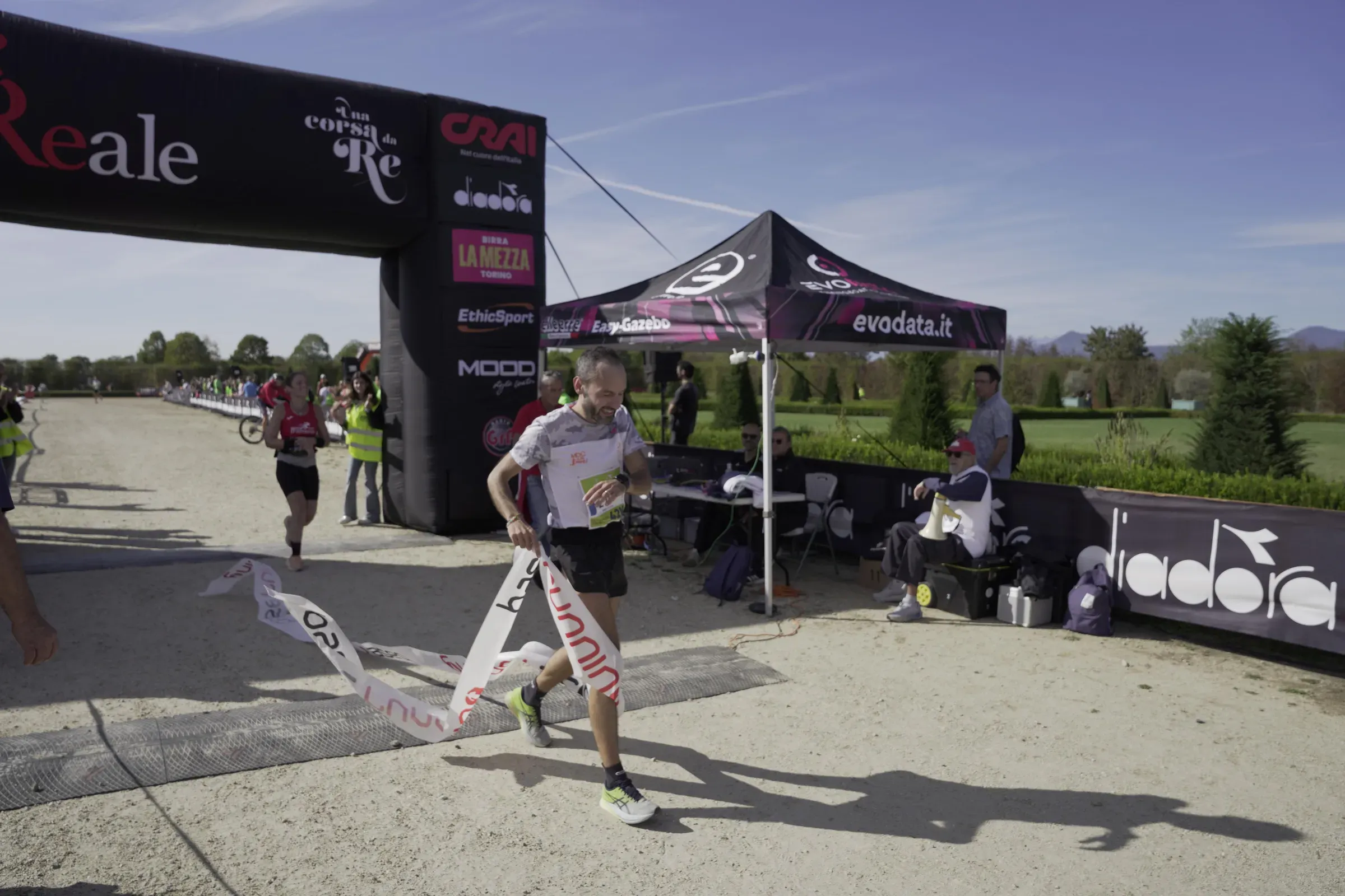 Un runner sorridente si porta dietro il nastro Base Running dopo il traguardo, sotto il cielo azzurro di Venaria
