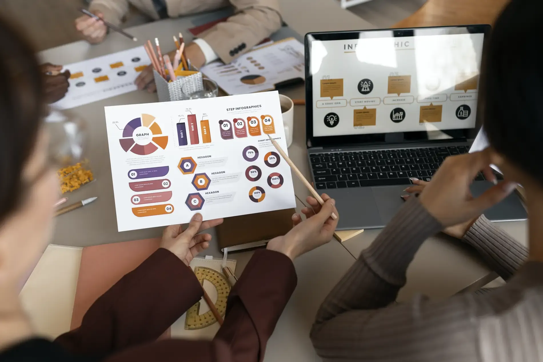 People at a work table analysing charts and infographics printed on a sheet of paper, next to a laptop showing more charts