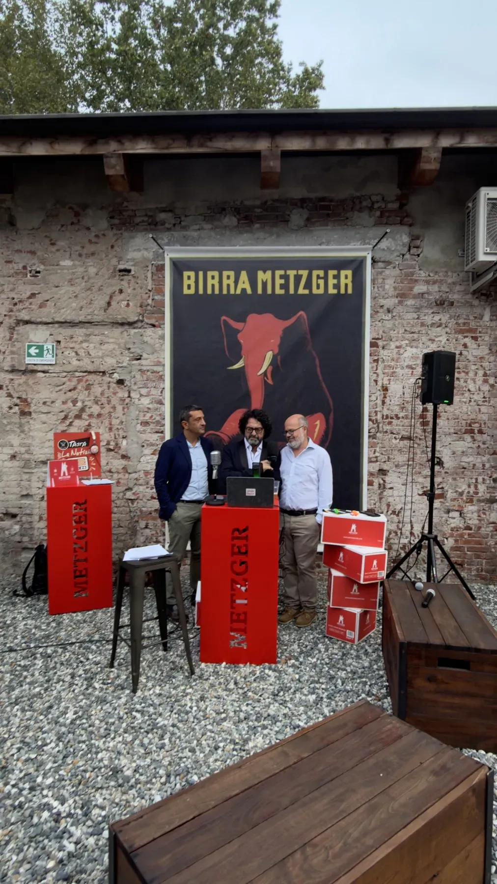 Un talk all'aperto al Metzger Fest, davanti al muro di mattoni dello stabilimento e al manifesto dell'elefante Birra Metzger, con i podi rossi Metzger e i microfoni