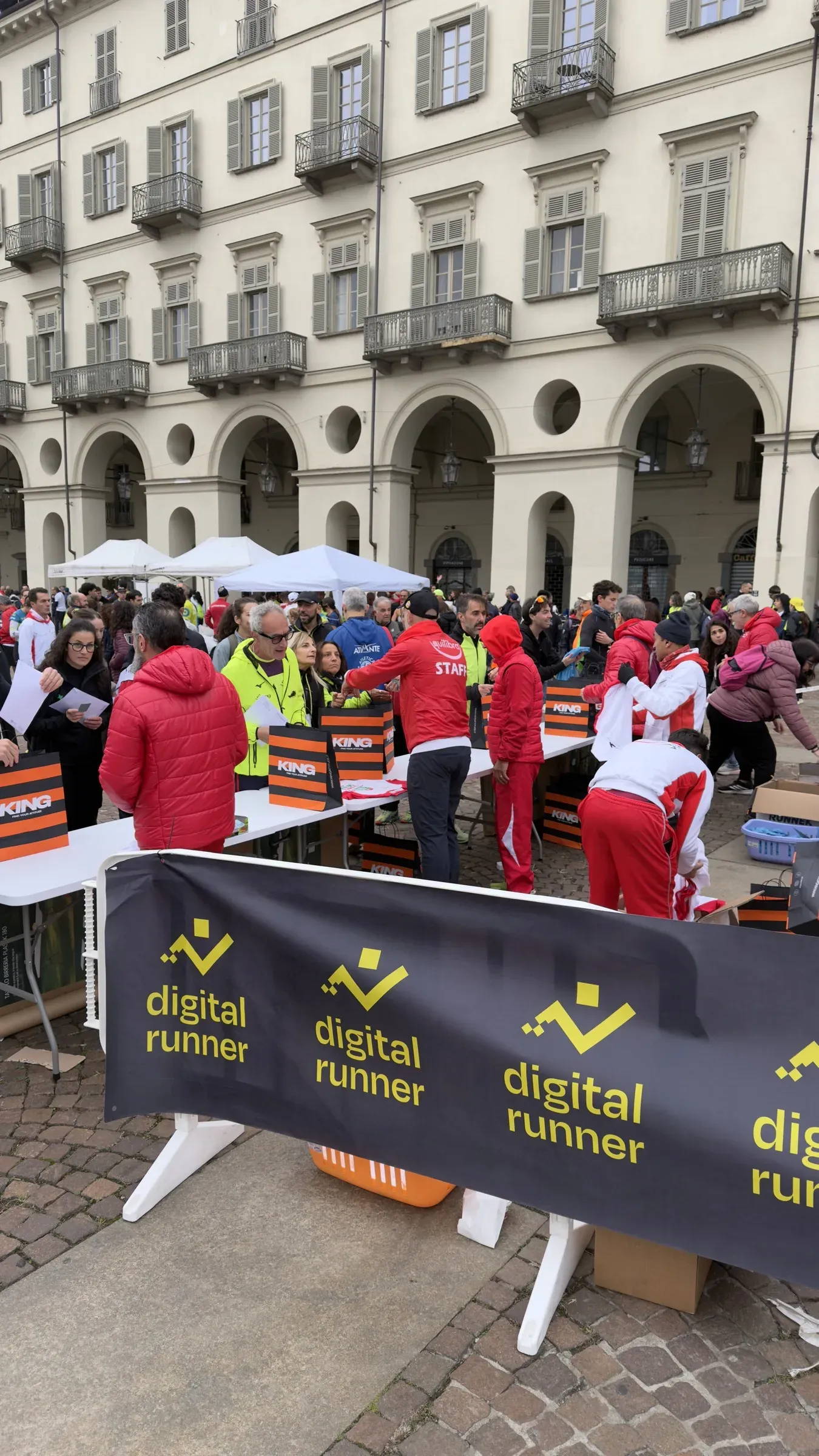 I volontari di Equilibra Running Team in rosso consegnano i pacchi gara sotto i portici di piazza Vittorio, dietro la transenna Digital Runner