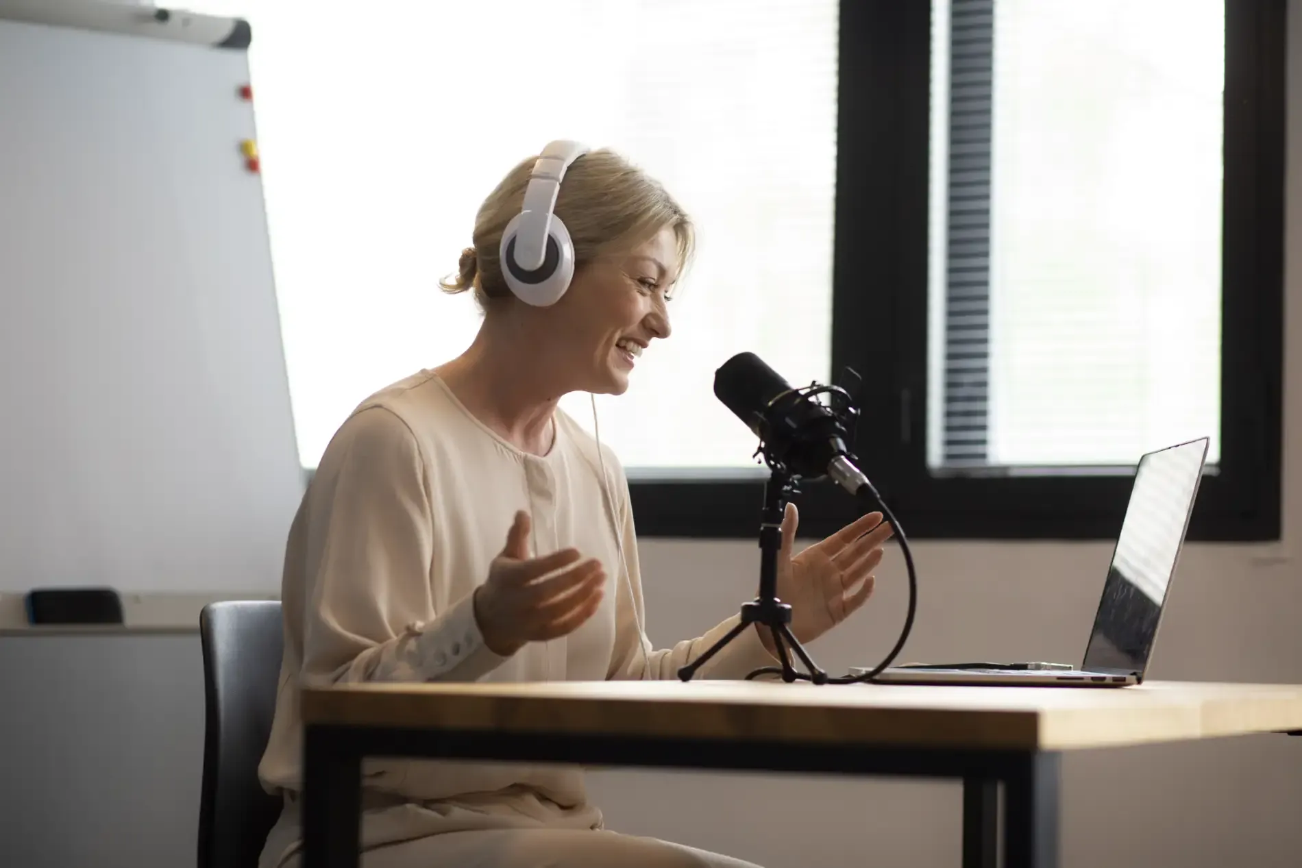 Donna sorridente con cuffie bianche che parla a un microfono da podcast davanti a un laptop, in un ufficio luminoso