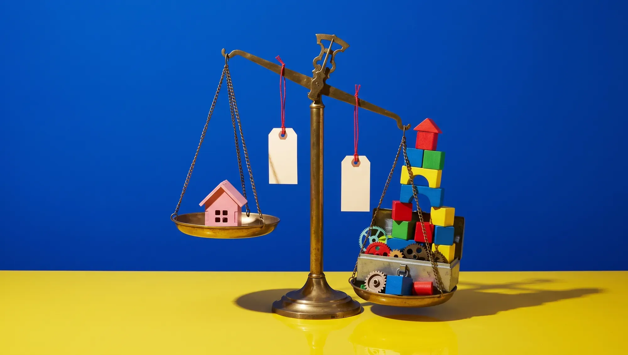 Sur un décor bleu et jaune, une vieille balance en laiton : sur un plateau une petite maison rose, sur l'autre une pile de jeux de construction, d'engrenages et d'outils, avec deux étiquettes de prix vierges