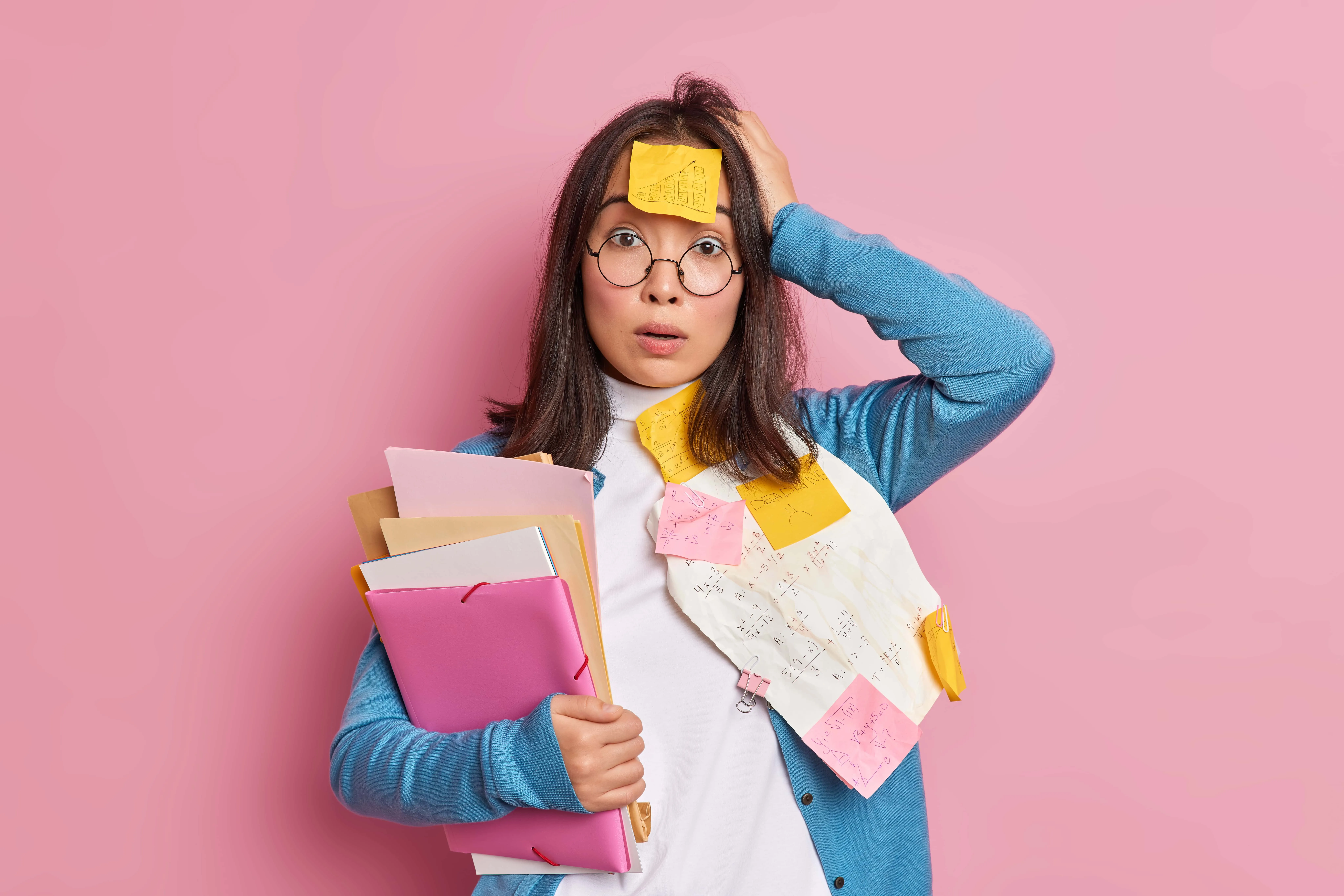 Ragazza con occhiali tondi e aria confusa, coperta di post-it e appunti, che stringe delle cartelline su sfondo rosa