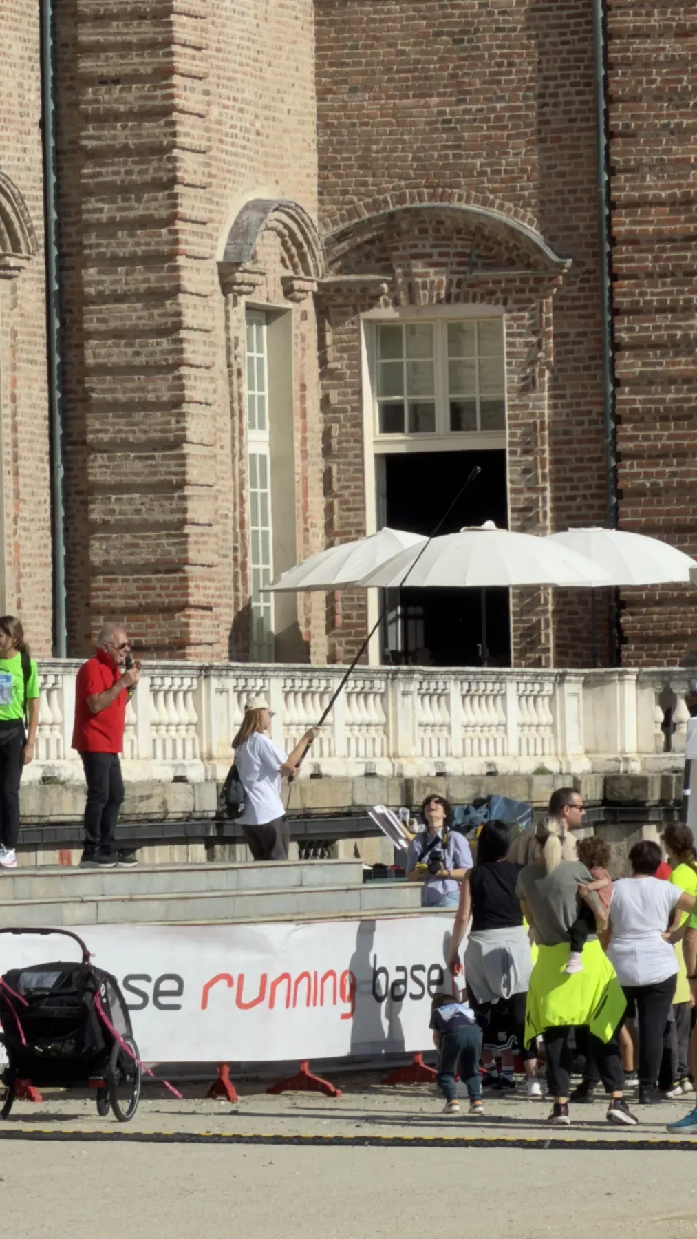 Dietro le quinte sul palco del Weekend Reale: lo speaker al microfono, una videomaker con l'asta e una fotografa davanti alla facciata in mattoni della Reggia di Venaria