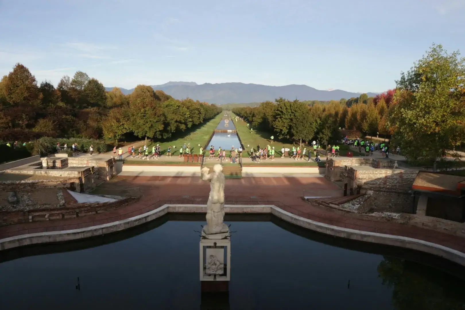 Il canale della Fontana d'Ercole dalla Reggia di Venaria: la statua in primo piano, i runner che attraversano i Giardini, gli alberi d'autunno e le montagne