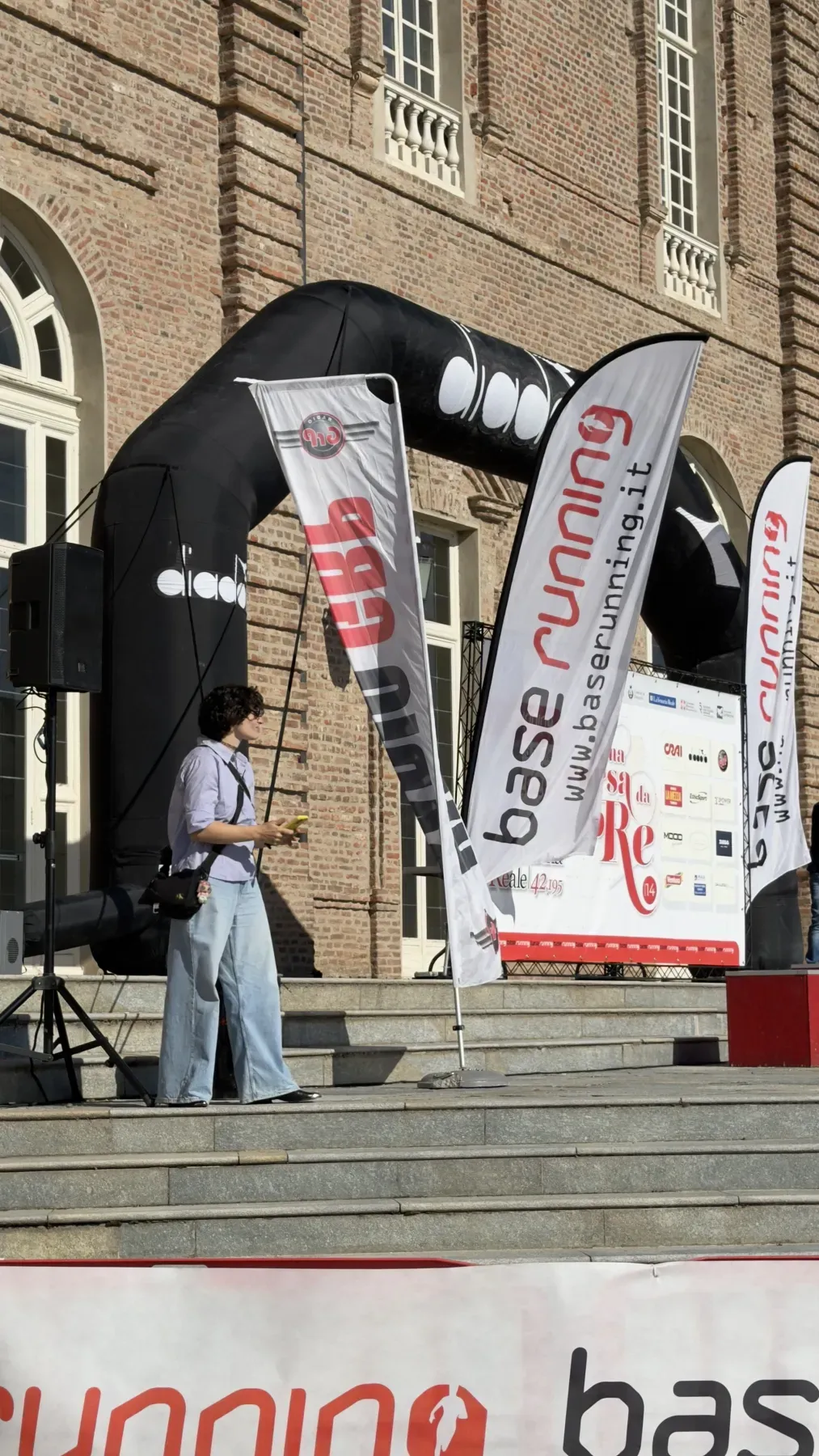 Una persona di Digital Runner sulla scalinata della Reggia di Venaria, tra le bandiere Base Running, l'arco Diadora e il pannello sponsor di Una Corsa da Re